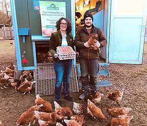 Hoteliére Frau Schmitt freut sich zusammen mit Landwirt Doppelhammer über Zusammenarbeit. Eier in den Händen. Im Vordergrund die Hühner. 